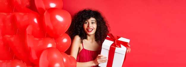 Vacances et célébration belle femme aux cheveux bouclés debout près de ballons coeur holding gift b