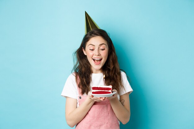 Vacances et célébration. Femme excitée célébrant son anniversaire, soufflant une bougie sur un gâteau, portant un gâteau de fête et s'amusant, debout sur fond bleu.