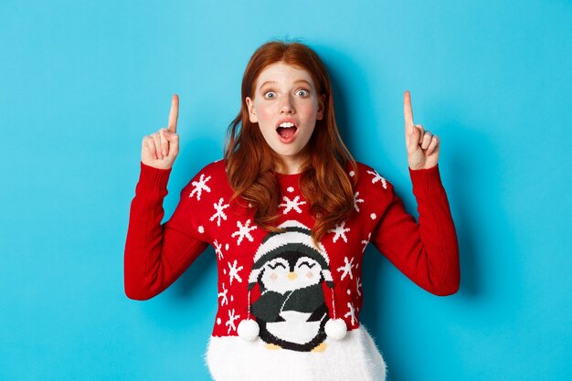 Vacances d'hiver et concept de célébration. Fille rousse étonnée montrant une publicité, pointant vers le logo de Noël, debout excitée sur fond bleu.