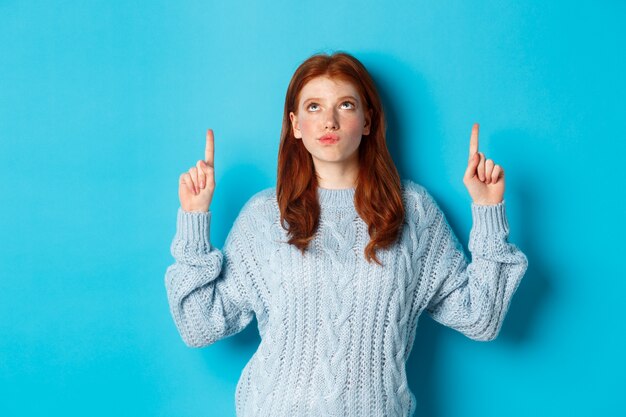 Vacances d'hiver et concept de personnes. Rousse réfléchie en pull regardant et pointant les doigts vers le haut, ayant des doutes, pensant ou faisant un choix, debout sur fond bleu