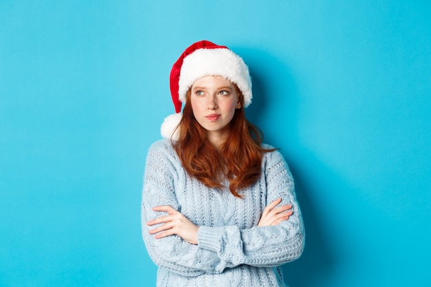 Vacances d'hiver et concept de réveillon de Noël. Femme rousse réfléchie en bonnet de Noel et pull, regardant à gauche et méditant, faisant des plans de Noël, debout sur fond bleu.