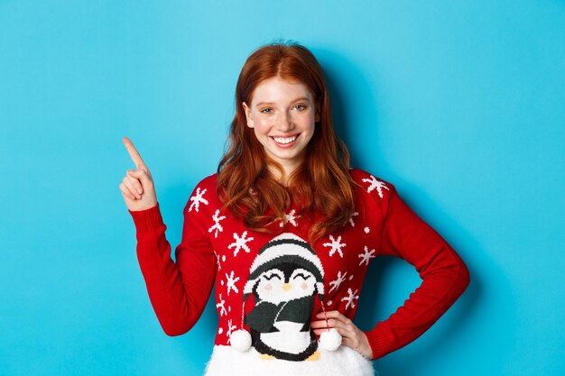 Vacances d'hiver et concept de réveillon de Noël. Jolie adolescente aux cheveux ondulés rouges, pointant vers le coin supérieur gauche et souriant à la caméra, debout sur fond bleu.