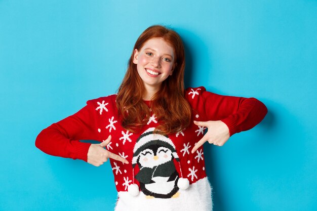 Vacances d'hiver et concept de réveillon de Noël. Jolie fille rousse pointant du doigt un joli pull de Noël avec un pingouin, debout sur fond bleu.