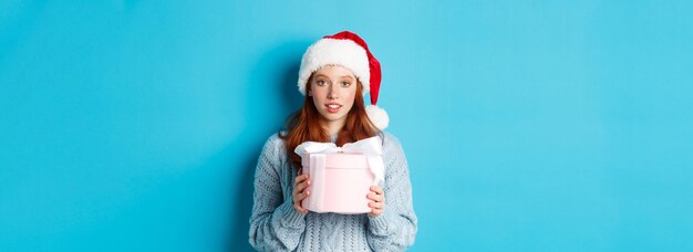 Vacances d'hiver et concept de réveillon de noël jolie fille rousse portant un bonnet de noel tenant un cadeau de nouvel an