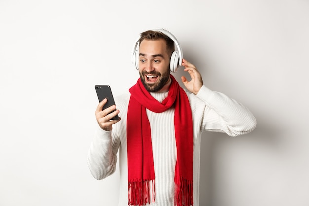 Vacances d'hiver et concept technologique. Heureux homme écoutant de la musique dans des écouteurs, regardant émerveillé par l'écran du mobile, debout sur fond blanc.