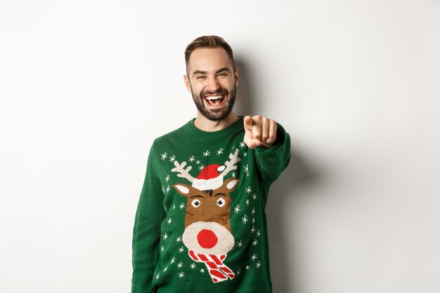 Vacances d'hiver et Noël. Heureux jeune homme célébrant le nouvel an, pointant le doigt vers vous et riant, debout sur fond blanc