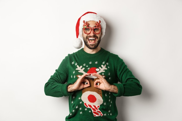 Vacances de Noël, concept de célébration. Heureux gars en bonnet de Noel et lunettes de fête se moquant d'un pull drôle, debout sur fond blanc.