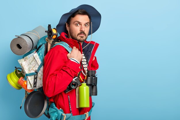 Le vacancier masculin a un repos actif, porte un sac à dos avec une carte, un chiffon enroulé, porte des vêtements touristiques décontractés, utilise des jumelles