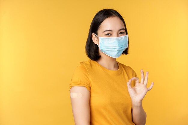 Vaccination contre le covid et le concept de santé Heureuse fille asiatique montrant bien portant un masque médical pansement sur l'épaule a reçu le vaccin contre le coronavirus tiré sur fond jaune