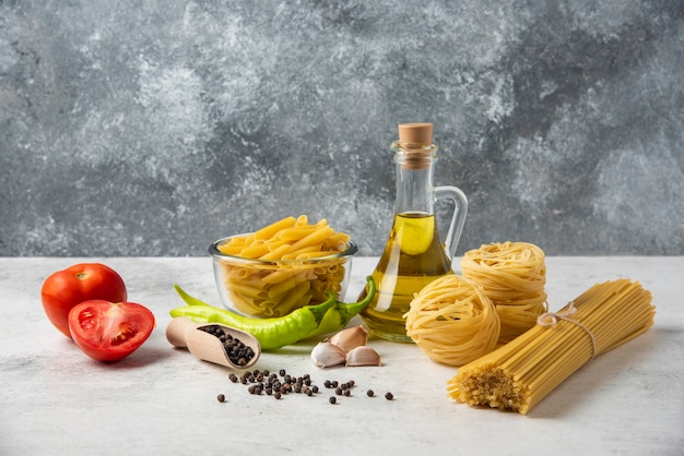 Variété de pâtes crues, bouteille d'huile d'olive, grains de poivre et légumes sur tableau blanc.