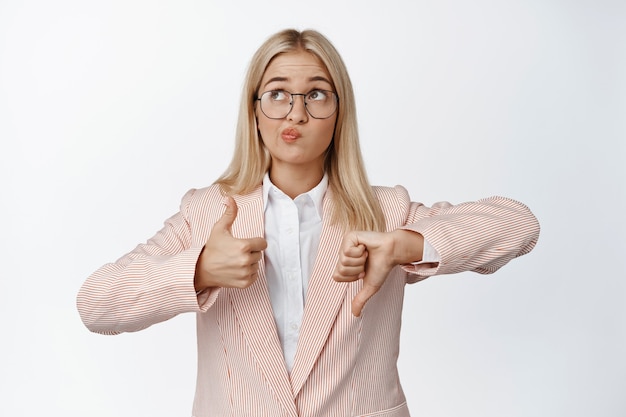 Vendeuse prenant une décision, montrant les pouces vers le haut et vers le bas, pensant comment évaluer, debout sur blanc.