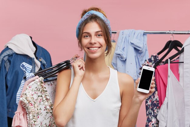 Vente au détail, vente, consommation et concept technologique moderne. Portrait de charmante jeune femme debout au rack avec des vêtements à la mode, profitant du shopping au centre commercial, payant avec application en ligne sur téléphone portable