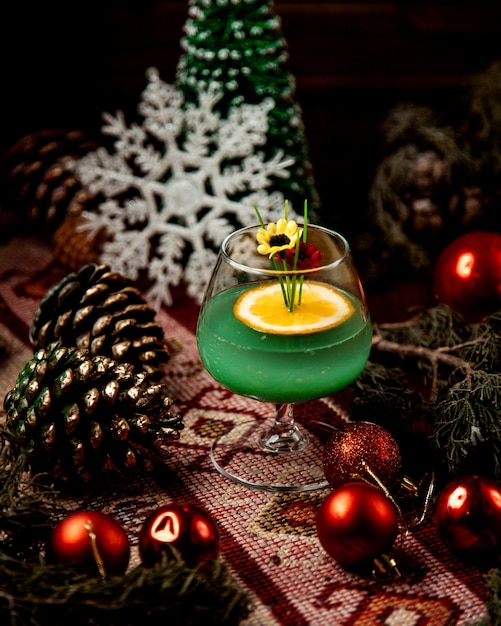 Un verre de boisson verte garnie de tranche d'orange et de fausses fleurs autour des décorations de Noël