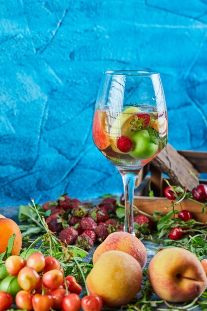 Un verre de jus de fruits, boîte de cerises, fraises fraîches et pêches sur surface bleue