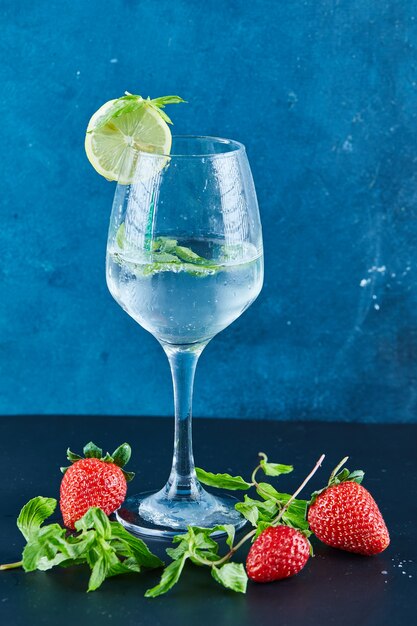 Un verre de jus à la menthe et une tranche de citron sur une surface bleue avec des fraises fraîches et de la menthe