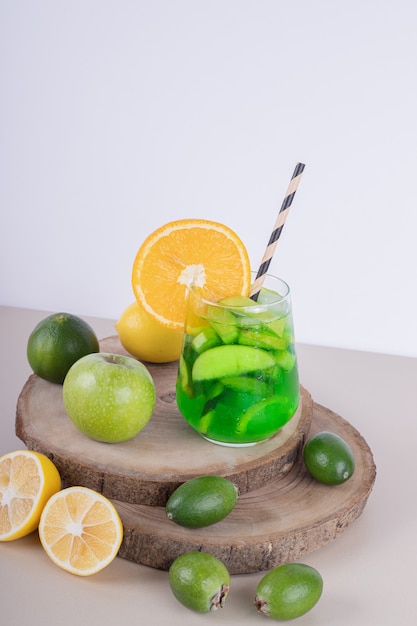 Un verre de jus avec des tranches de fruits et des fruits frais sur un mur blanc.