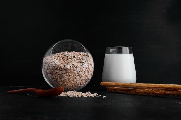 Un verre de lait servi avec une tasse de grains sur une surface noire.