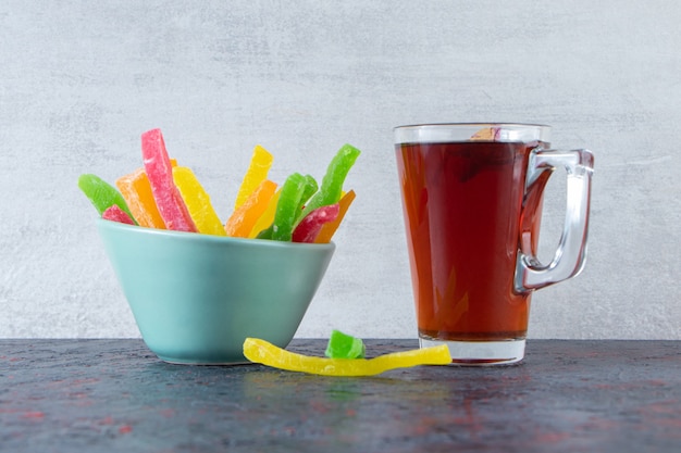 Verre de thé noir avec bol de bonbons colorés sur une surface sombre.