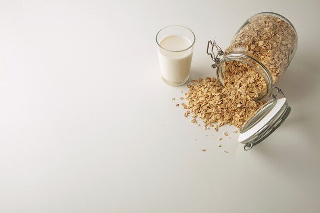 Verre transparent avec du lait biologique frais près de couché pot rustique ouvert avec des flocons d'avoine répartis isolé au centre sur la vue de côté de dessus de table blanche