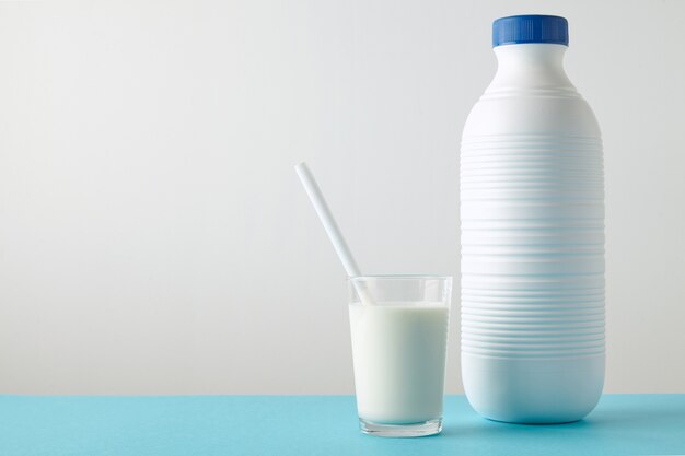 Verre transparent avec du lait frais et de la paille blanche à l'intérieur près d'une bouteille en plastique à rayures vierge avec bouchon bleu