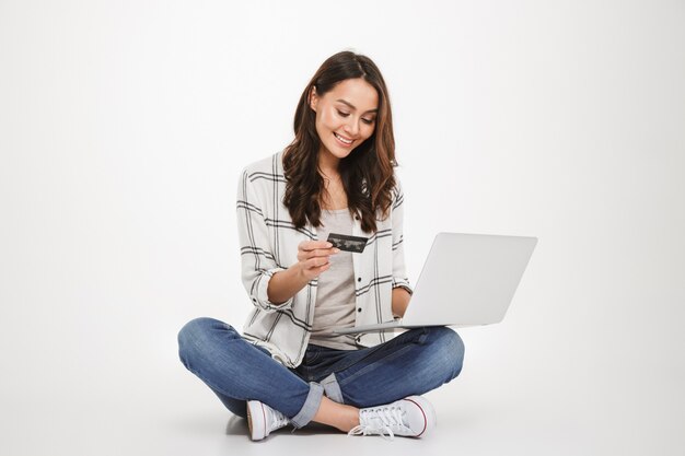 Vertical femme brune heureuse en chemise assise sur le sol avec ordinateur portable et carte de crédit sur gris