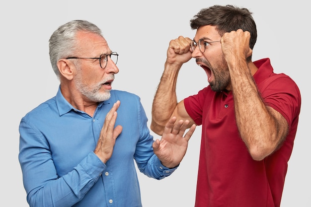 Vexé irrité père et jeune fils adulte posant contre le mur blanc