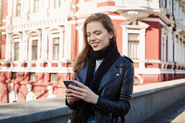 Vie des grandes villes. Portrait de charmante femme élégante, debout dans la rue, tenant un smartphone et des SMS