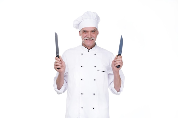 Vieil homme, chef professionnel en uniforme tenant deux couteaux en métal tout en restant sur un mur blanc