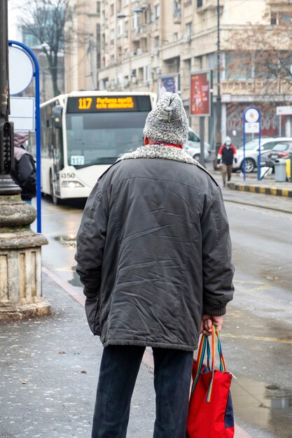 Un vieil homme dans une veste et un chapeau en laine sur un arrêt, rue sur l'arrière-plan, temps nuageux à Bucarest, Roumanie
