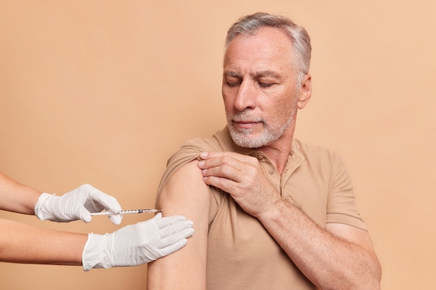 Un vieil homme sérieux aux cheveux gris reçoit une injection de vaccin contre le coronavirus porte un t-shirt isolé sur un mur de studio beige