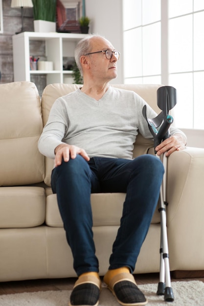 Un vieil homme solitaire assis sur un canapé dans une maison de retraite tenant des béquilles et regardant ailleurs.