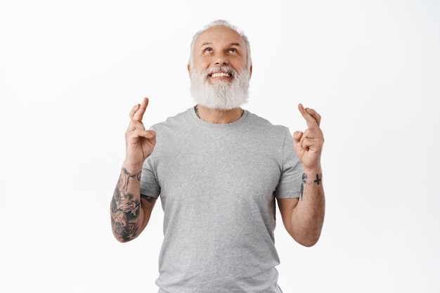 Vieil homme souriant avec des tatouages levant les yeux dans le ciel, fait un vœu et croise les doigts pour la bonne chance, priant, suppliant Dieu pour la fortune ou le rêve devenu réalité, debout contre un mur blanc