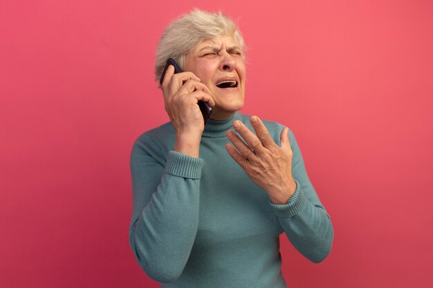 Vieille femme agacée portant un pull à col roulé bleu parlant au téléphone en gardant la main en l'air avec les yeux fermés isolé sur un mur rose avec espace pour copie