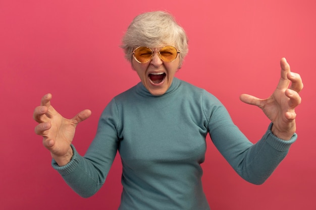 Photo gratuite vieille femme en colère portant un pull à col roulé bleu et des lunettes de soleil gardant les mains en l'air en criant les yeux fermés