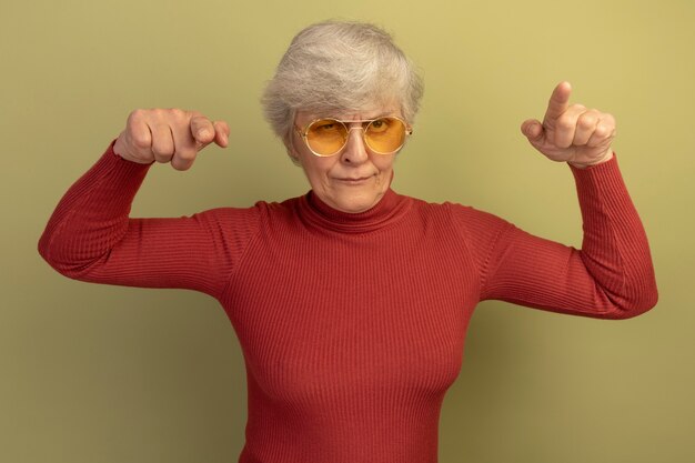 Vieille femme confiante portant un pull à col roulé rouge et des lunettes de soleil regardant et pointant vers l'avant isolé sur un mur vert olive