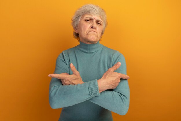 Vieille femme confuse portant un pull à col roulé bleu gardant les mains croisées pointant sur les côtés isolés sur un mur orange avec espace de copie