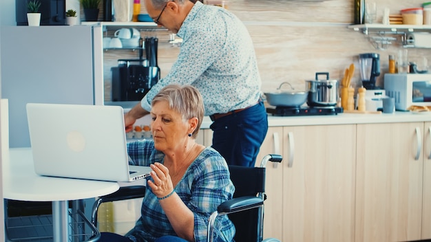 Vieille femme handicapée en fauteuil roulant travaillant sur ordinateur portable dans la cuisine. Vieille personne âgée handicapée paralysée utilisant la technologie de communication Internet en ligne moderne.