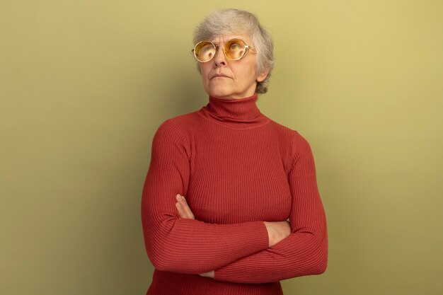 Une vieille femme réfléchie portant un pull à col roulé rouge et des lunettes de soleil debout avec une posture fermée levant isolée sur un mur vert olive avec espace pour copie
