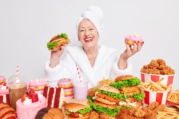 Une vieille femme souriante et gaie se sent très heureuse tient un délicieux hamburger et un beignet portant un peignoir et une serviette sur la tête mange de la malbouffe