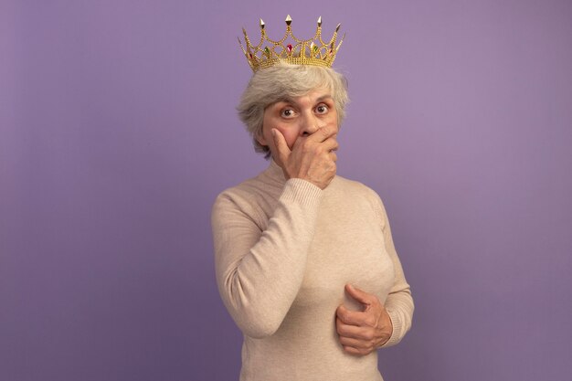 Une vieille femme surprise portant un pull à col roulé crémeux et une couronne mettant la main sur la bouche et sur le ventre