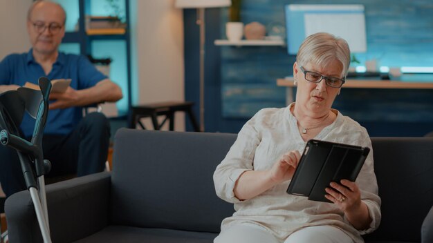 Vieille femme utilisant une tablette numérique avec écran tactile dans le salon. Personne âgée avec des béquilles sur un canapé naviguant sur Internet sur un appareil pour s'amuser et profiter de la retraite à la maison. Personne moderne