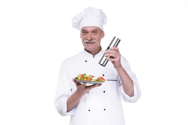 Photo gratuite vieux cuisinier masculin tenant une assiette avec salade de légumes et poivre sur mur blanc