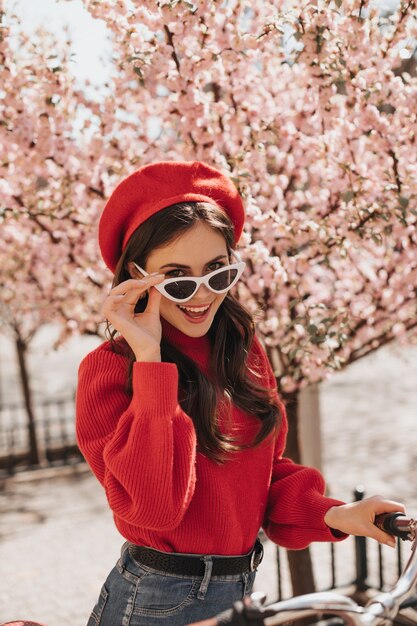 Vilaine fille aux cheveux ondulés enlève ses lunettes et se penche sur la caméra. Jolie femme brune en béret, pull rouge et jeans posant avec vélo contre sakura