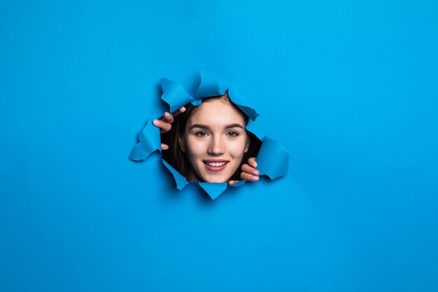 Visage de jolie jeune femme regardant à travers le trou bleu dans le mur de papier.