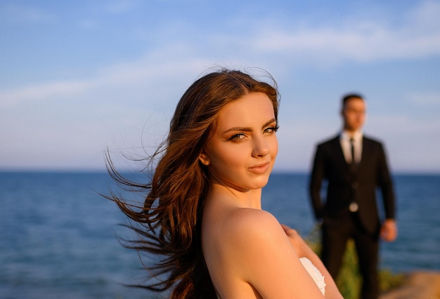 Visage de portrait de jolie mariée en robe de mariée à manches ouvertes tandis que marié élégant derrière debout sur fond de paysage de mer Soleil brillant sur le visage attrayant de la femme avec le maquillage de mariage Nature