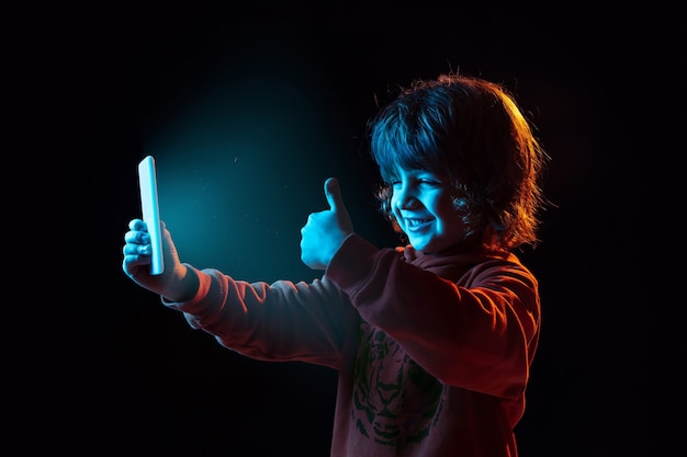 Vlogging avec smartphone, pouce vers le haut. Portrait de garçon caucasien sur un mur sombre en néon. Beau modèle bouclé. Concept d'émotions humaines, expression faciale, ventes, publicité, technologie moderne, gadgets.