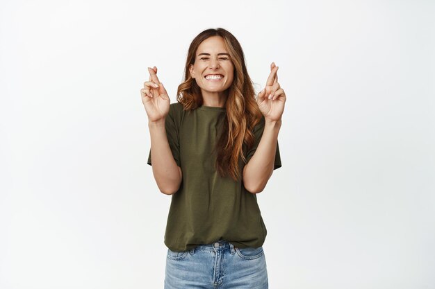 Vœux. Une femme adulte souriante croise les doigts, ferme les yeux, pense positivement, souhaite que le rêve devienne réalité, espère atteindre le prix de l'objectif, debout excitée sur fond blanc.