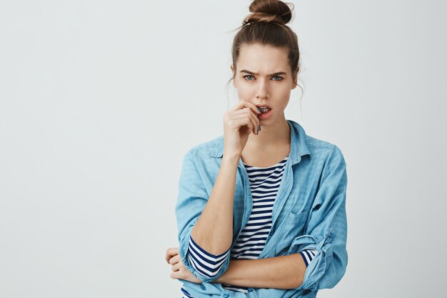 Vos mots ne correspondent pas à la vérité. Portrait de jolie jeune femme avec coiffure chignon, fronçant les sourcils et mordant le pouce tout en regardant avec incrédulité, debout