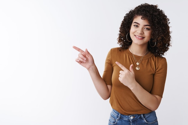 Vous devriez vérifier. Belle jeune fille souriante et gaie utile pointant l'index gauche souriant joyeusement montrant le meilleur choix de promotion du produit indiquant l'espace de copie vierge, fond blanc