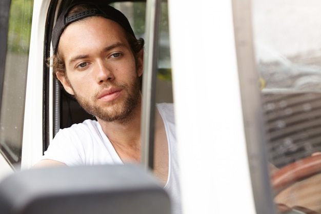 Voyage, aventure et concept de mode de vie actif. Élégant jeune homme en snapback assis à l'intérieur de son véhicule blanc